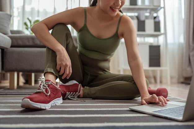 Eine sportliche Frau in Sportkleidung sitzt mit Hanteln auf dem Boden und benutzt zu Hause einen Laptop