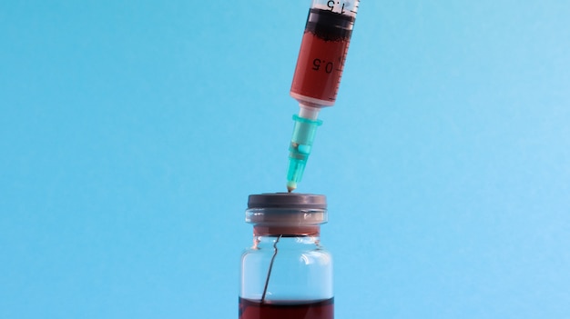 Eine Spritze ragt aus einer roten Flüssigkeitsflasche heraus. Auf blauem Hintergrund isoliert. Medizin, Injektionen, Impfstoffe und Einwegspritzen, Medikamentenkonzept. Sterile Flasche. Medizinische Glasampulle zur Injektion.