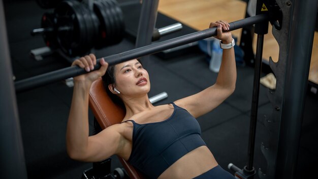 Eine starke asiatische Frau in Sportkleidung hebt eine Stange auf einer Bankpresse und trainiert in einem Fitnessstudio