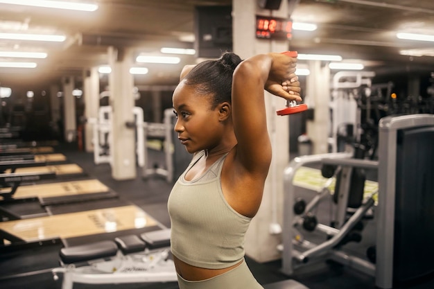 Eine starke schwarze Sportlerin übt im Fitnessstudio eine Trizepsverlängerung mit Hanteln.