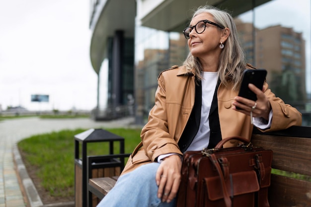 Eine stilvolle ältere Frau mit einem Handy in den Händen ruht auf einer Bank und schaut sich um