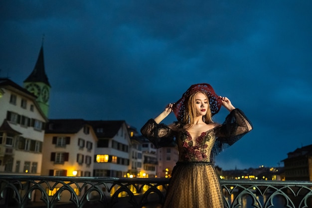 Eine stilvolle Braut in einem schwarzen Hochzeitskleid und einem roten Hut posiert nachts in der Altstadt von Zürich. Schweiz.