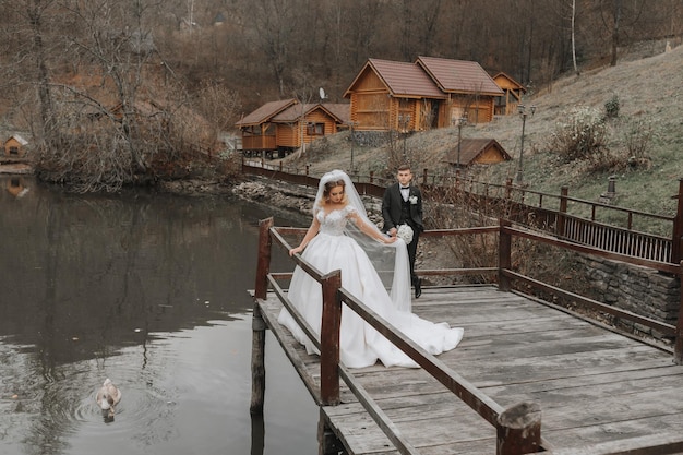 Foto eine stilvolle braut in einem üppigen kleid und modischer frisur steht auf einem pier in einem park in der nähe von holzhäusern der bräutigam hinter ihr schwäne schwimmen im see