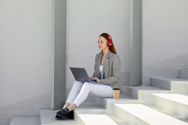 Eine stilvolle Frau arbeitet im Freien und hört Musik, während sie auf der Treppe sitzt. Möglichkeit, überall zu arbeiten