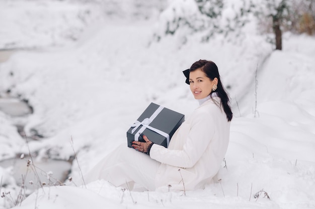Eine stilvolle Frau mit einem weißen Anzug mit einem Neujahrsgeschenk in ihren Händen in einem Winterwald Ein Mädchen in der Natur in einem verschneiten Wald mit einer Geschenkbox