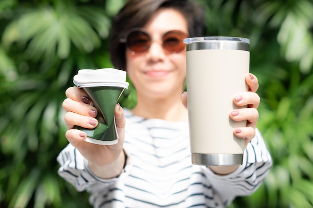 Eine stilvolle, schöne Frau, die eine Kaffeetasse zum Mitnehmen in beiden Händen hält. Eine ist eine Einweg-Pappbecher mit Plastikdeckel, die andere ist ein wiederverwendbarer Edelstahlbecher. Kein Stroh- und Null-Abfall-Konzept.