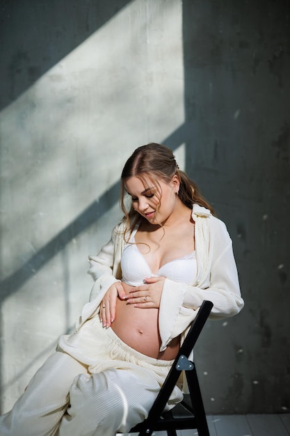 Foto eine stilvolle schwangere junge frau in einem weißen anzug sitzt auf einem stuhl und berührt ihren bauch mit einem sanften lächeln. sie wartet auf die geburt, fürsorge und mutterschaft, liebe und zärtlichkeit