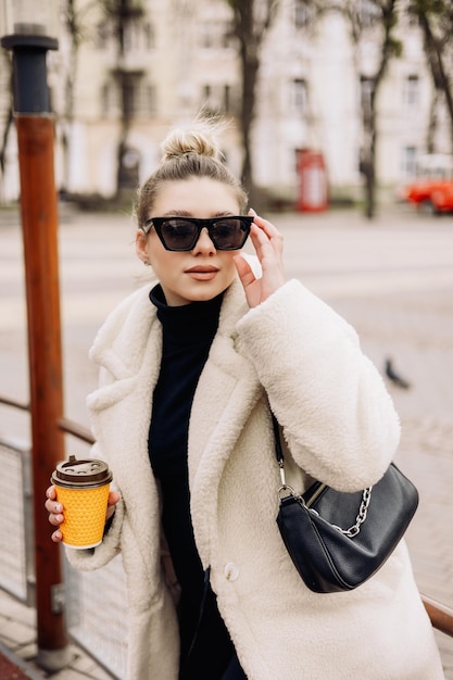 Eine süße junge Frau in einem weißen Pelzmantel und einer Tasse Kaffee geht durch die Stadt Stilvolle junge schwangere Mädchen Casual Fashion für Damen