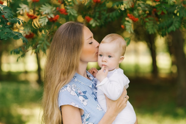 Eine süße und sanfte junge Frau, eine europäische Mutter, hält ihren kleinen Sohn in den Armen und küsst ihn mit roter Eberesche in der Nähe der Zweige