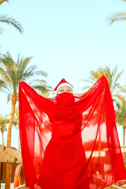 Eine Tänzerin eines orientalischen Tanzes in einer Weihnachtsmütze vor einem Hintergrund von Palmen mit geschlossenem Umhang, ihr Gesicht, nur ihre Augen sind sichtbar