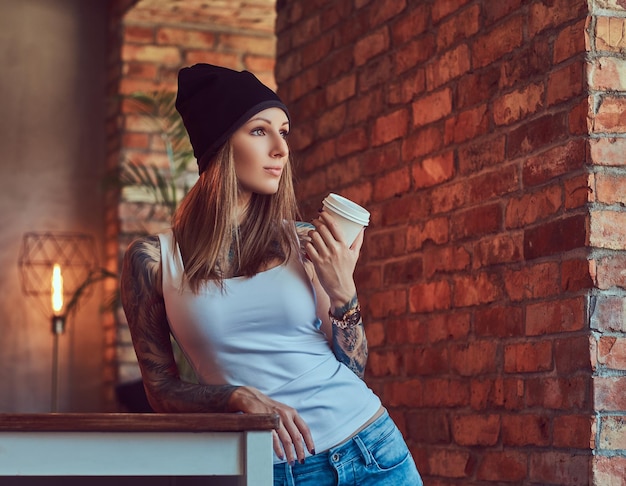 Eine tätowierte sexy Blondine in einem T-Shirt und einem Hut mit einer Tasse Kaffee in einem Raum mit Loft-Interieur.