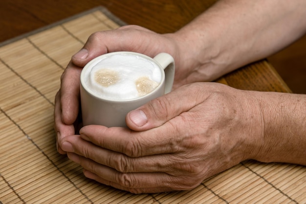 Eine Tasse frischen Kaffee mit Cappuccino-Schaum in den Händen eines Mannes in der Nähe