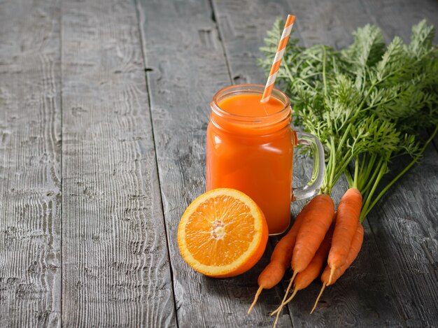 Eine Tasse frischen Karotten-Smoothie mit Cocktailstroh, Karotten und Orangen auf einem rustikalen Tisch.
