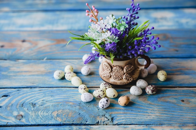 Eine Tasse frühlingspurpurne Blumen und eine Streuung von Wachteleiern auf einem alten, abgenutzten blauen Tisch.