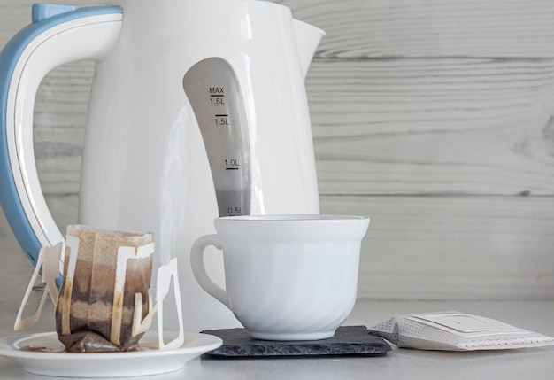 Eine Tasse gebrühter natürlicher Kaffee aus einem Tropfbeutel auf dem Hintergrund einer Teekanne in der Nähe