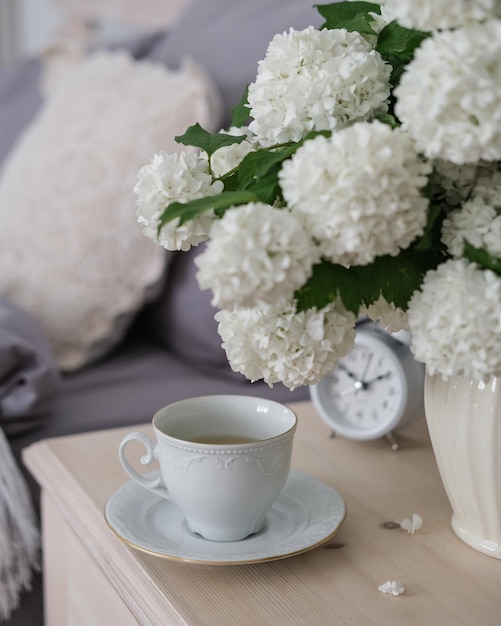 Eine Tasse Kaffee, ein Wecker und eine Vase mit weißen Blumen auf dem Nachttisch. Teezeit