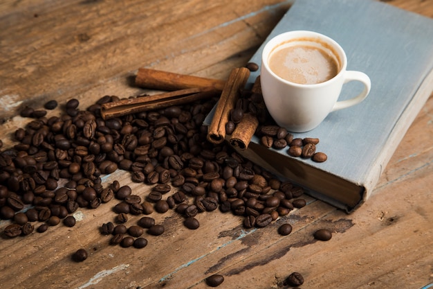 Eine Tasse Kaffee, Kaffeesamen, Zimt und ein Buch auf einem Holztisch