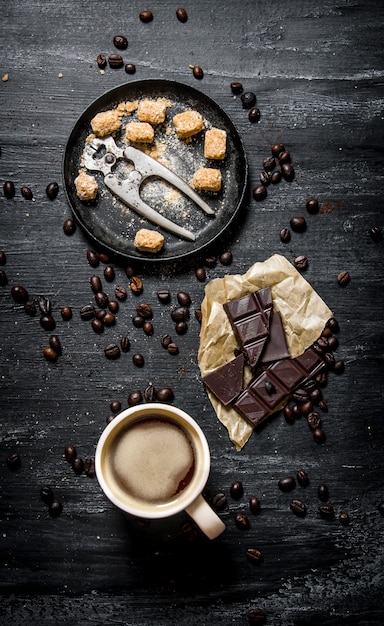 Eine Tasse Kaffee mit Bitterschokolade und dunkelbraunem Zucker. Auf schwarzem rustikalem Hintergrund.