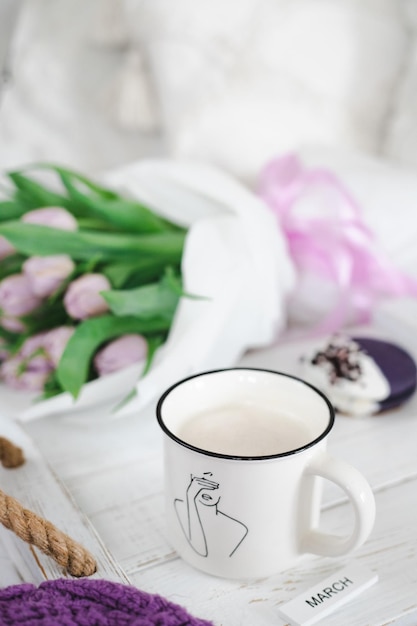 Eine Tasse Kaffee mit einer Katze darauf und ein Strauß Tulpen auf einem weißen Tisch Inschrift März