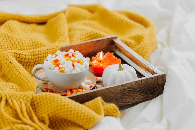 Eine Tasse Kaffee mit Marshmallow und Zuckermais auf dem Bett mit warmem Plaid. Herbstgetränk mit Kerze und Kürbis auf Holztablett. Hygge-Konzept.