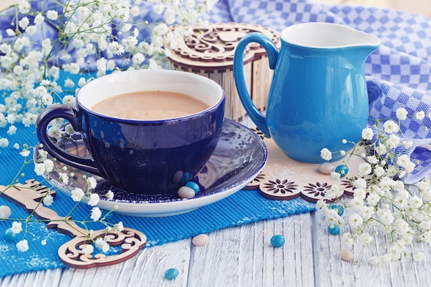 Eine Tasse Kaffee mit Milch wird von einer blauen Serviette, einem Holzschlüssel und kleinen weißen Blumen (Gypsophila) auf einem weißen Holztisch dekoriert