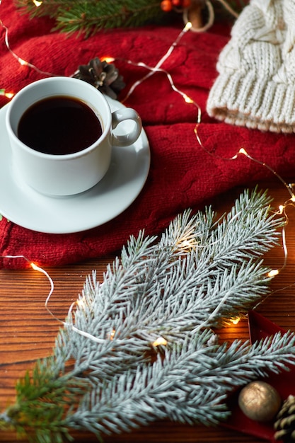 Eine Tasse Kaffee und eine Girlande auf einem Holztablett mit einem gemütlichen roten Pullover. Neujahrsstillleben mit Tannenzweig und Eberesche