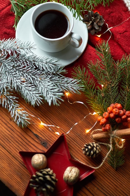 Eine Tasse Kaffee und eine Girlande auf einem Holztablett mit einem gemütlichen roten Pullover und einem warmen Hut. Neujahrsstillleben mit Tannenzweig