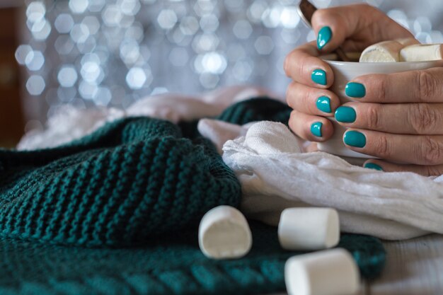 Eine Tasse Kakao mit Marshmallow in weiblichen Händen mit blauer Maniküre auf glänzendem Hintergrund