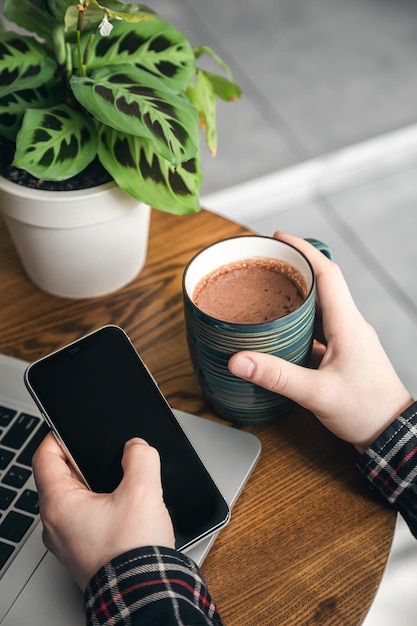 Eine Tasse Kakao und ein Smartphone in den Händen eines Mannes vor einem Laptop