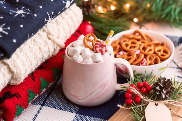 Eine Tasse mit Marshmallow in der Nähe eines Stapels Winterpullover eine Schüssel mit Brezeln Weihnachtsschmuck