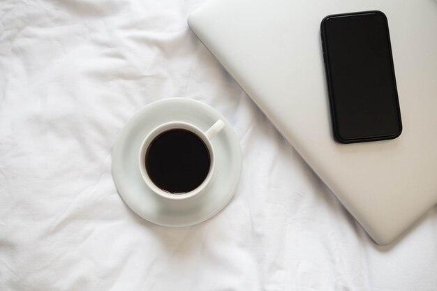 Eine Tasse schwarzer Kaffee, ein Laptop und ein Smartphone auf einem unfertigen weißen Bett.