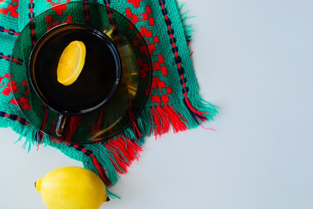 Eine Tasse Tee mit Zitrone steht auf einer dunklen Untertasse, die daneben auf einem gestrickten grünroten Schal steht ...
