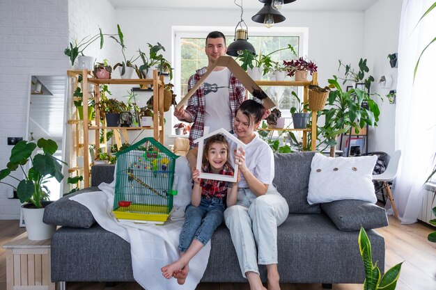 Foto eine traditionelle familie mit einem kind und einem papageienhaustier in einem käfig genießt ein neues zuhause, sitzt auf dem sofa und hält das dach fest. kauf einer hypothekenversicherung und einzug in das eigene zuhause