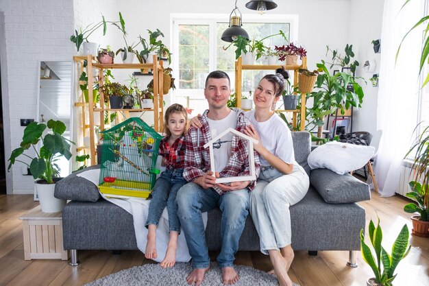 Eine traditionelle Familie mit einem Kind und einem Papageienhaustier in einem Käfig genießt ein neues Zuhause, sitzt auf dem Sofa und hält das Dach fest. Kauf einer Hypothekenversicherung und Einzug in das eigene Zuhause