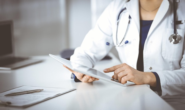 Eine unbekannte junge Ärztin überprüft einige Medikamentennamen, während sie in einer Klinik am Schreibtisch in ihrem Schrank sitzt. Ein Arzt mit einem Stethoskop benutzt einen Tablet-Computer, Close-up. Perfekte medizinische S