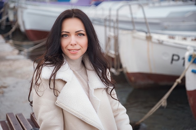 Eine unbeschwerte kaukasische Frau in beigefarbener Kleidung, die an einem warmen, windigen Tag den Blick auf das Meer genießt. Porträt vor einer Meeresbucht mit Booten am Pier