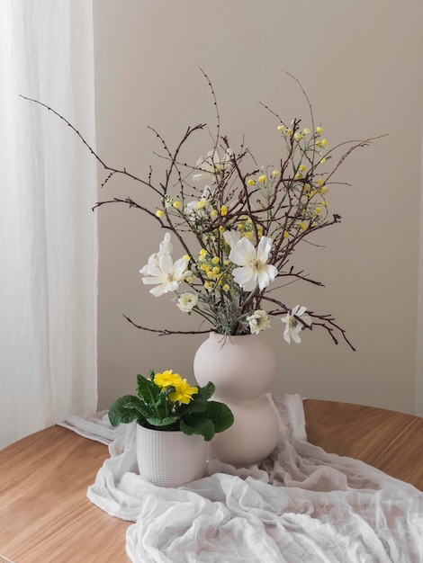 Eine Vase mit Blumen und eine hausgemachte Primroseblume in einem Topf auf einem runden Holztisch mit einem weißen Tischtuch