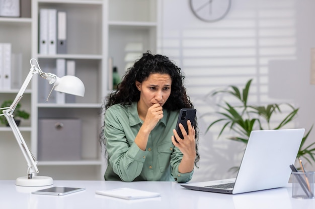 Eine verärgerte junge hispanische Frau sitzt im Büro am Schreibtisch und schaut besorgt auf das Telefon