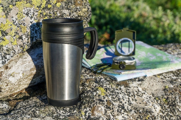 Eine Verschnaufpause im Weg - eine Karte, ein Kompass und ein Thermobecher auf einem Stein auf natürlichem Hintergrund