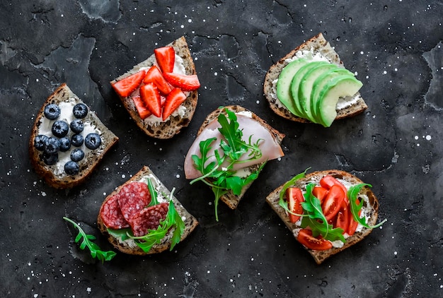 Eine Vielzahl von Frühstücks-Tapas-Snack-Sandwiches auf dunklem Hintergrund mit Avocado-Frischkäse-Schinken-Beeren-Salami-Kirschtomaten