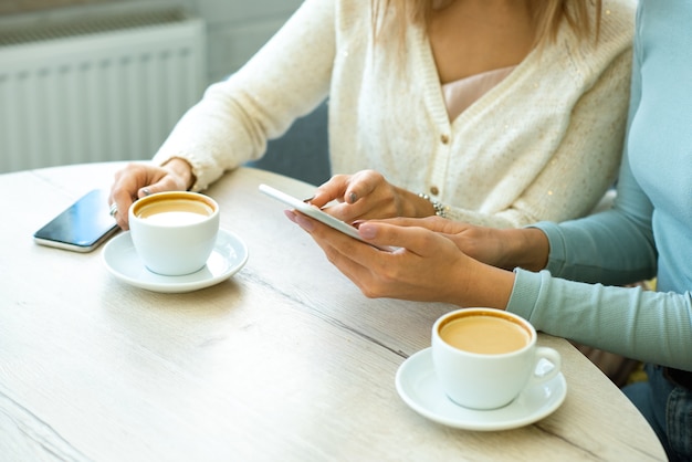Eine von zwei jungen zeitgenössischen Frauen, die auf Smartphonebildschirm zeigen, während sie Online-Waren mit Freund bei einer Tasse Kaffee im Café besprechen