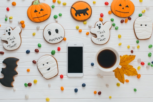 Foto eine vorbereitung für halloween: tasse kaffee und smartphone auf dem weißen hölzernen hintergrund mit süßigkeiten und lebkuchen.