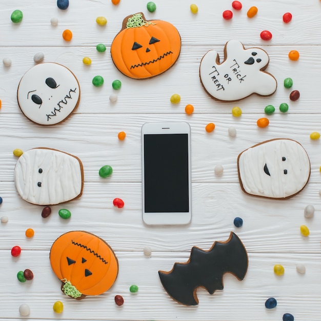 Eine Vorbereitung für Halloween: Tasse Kaffee und Smartphone auf dem weißen hölzernen Hintergrund mit Süßigkeiten und Lebkuchen.