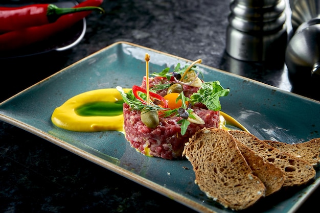 Eine Vorspeise vor dem Hauptgericht - Rindersteak-Tartar, serviert mit Croutons, Cappers, Eigelb, Gurken in einem blauen Teller auf einem Marmortisch. Restaurant Essen. Rohes Fleisch