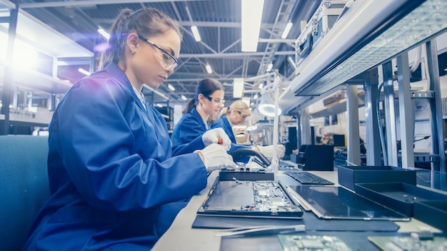 Foto eine weibliche elektronikfabrikarbeiterin in blauem arbeitsmantel und schutzbrille montiert laptop-mutterplatten mit einem schraubenzieher in einer hightech-fabrik mit mehreren angestellten.