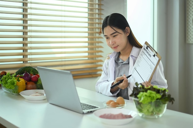 Eine weibliche Ernährungswissenschaftlerin zeigt vor dem Laptop das Clipboard und erklärt dem Patienten die Einzelheiten des Schlankheitsprogramms