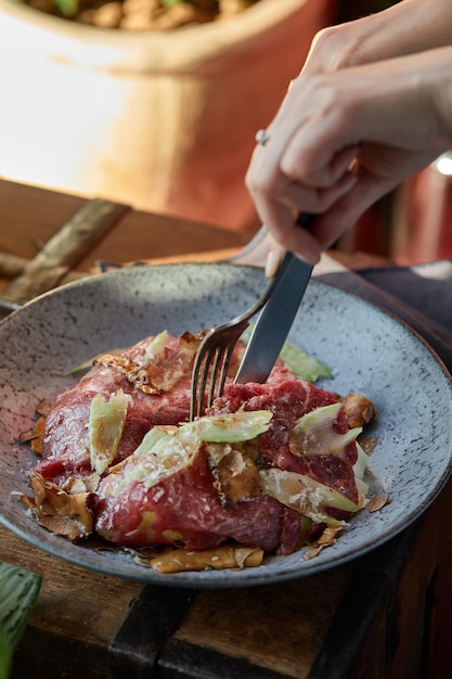 Eine weibliche Hand bereitet einen Gemüsesalat mit Rinderbraten zu. Ein Salatteller mit Rinderbraten, Zwiebeln, Rucola, Pfeffer und Tomaten auf einem schwarzen Tisch. Ansicht von oben.