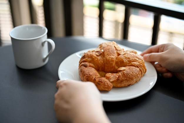 Eine weibliche Hand, die ein köstliches frisches französisches Croissant hält, serviert mit heißem Kaffee oder Kakao