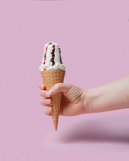 Eine weibliche Hand hält eine Eistüte mit Kirschmarmelade in einem Waffelglas auf rosafarbenem Hintergrund.