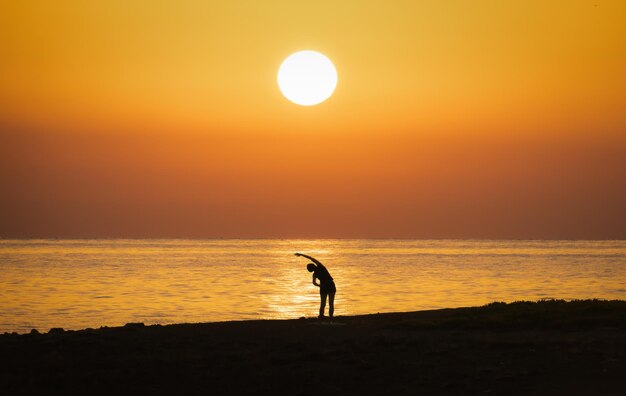 Eine weibliche Silhouette macht morgendliche Übungen am Ufer vor dem Hintergrund des Sonnenaufgangs